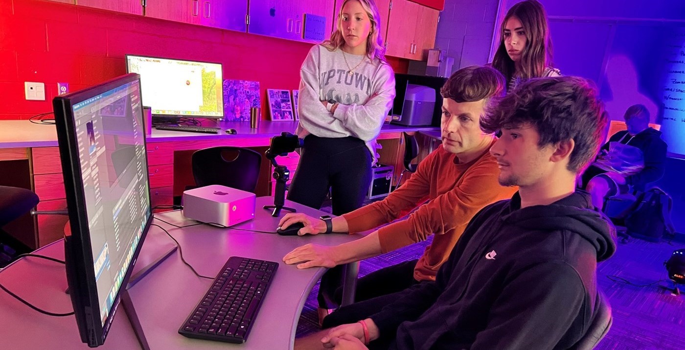 A teacher assisting a student with video editing, with 2 student watching.